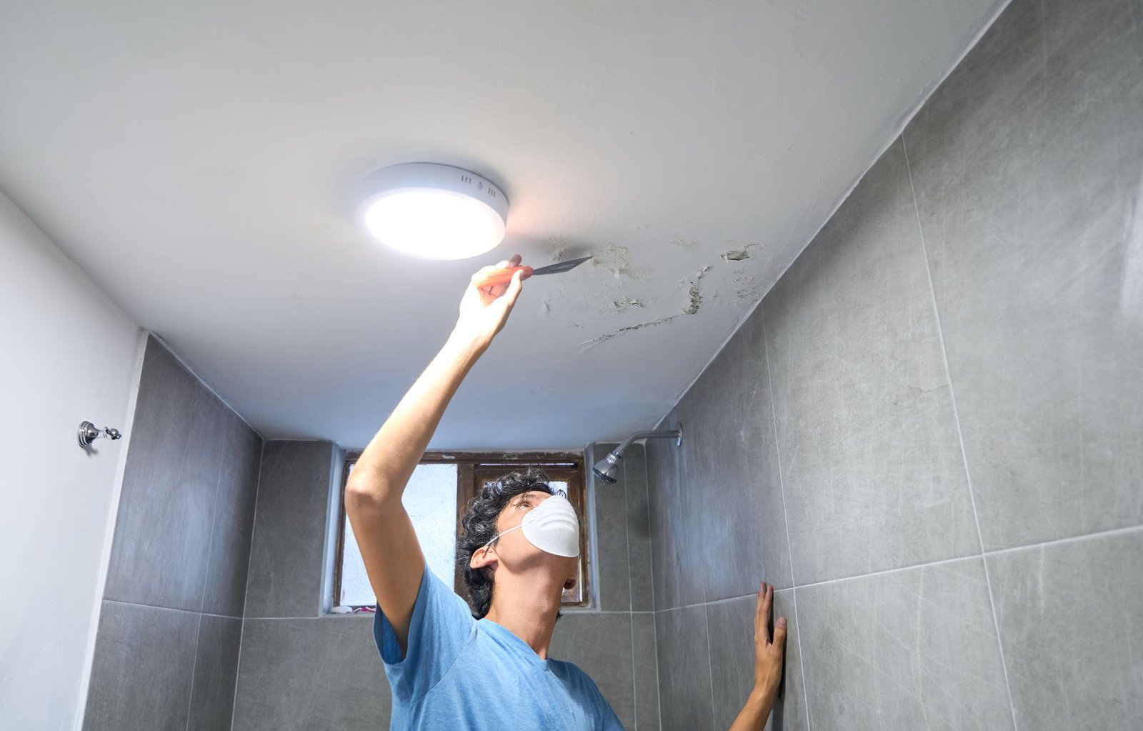 A man is adjusting a ceiling light fixture in a bathroom, emphasizing the role of upkeep in mold removal services in Honolulu, HI.